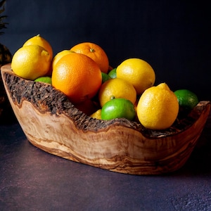 Puede incluir: Un cuenco de madera lleno de naranjas, limones y limas. El cuenco tiene un borde natural y rústico y está hecho de madera de color marrón claro. Las frutas son de colores brillantes sobre un fondo oscuro.