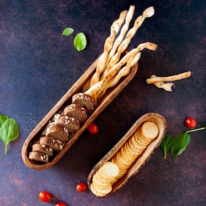 May include: Overhead shot of a wooden serving tray filled with breadsticks, sliced bread, and crackers. The tray is surrounded by fresh basil leaves and small red tomatoes on a dark, textured surface.