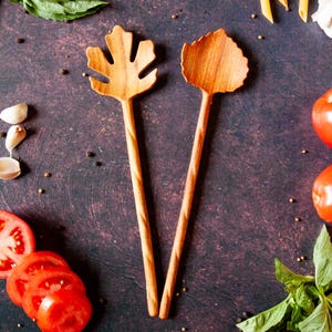May include: Two wooden salad servers with long, twisted handles. One server features a head shaped like a maple leaf with cutouts, while the other has a solid, rounded leaf-shaped head. Both utensils are crafted from light brown wood with a visible grain.