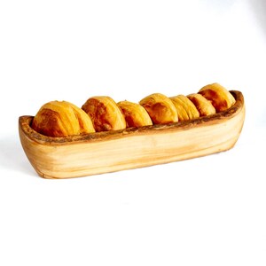 May include: A wooden bread bowl filled with golden-brown rolls. The bowl is carved from a single piece of light-colored wood, with a natural edge. The rolls are freshly baked and have a slightly crusty appearance, perfect for serving at a meal.