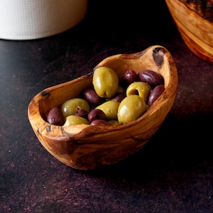 May include: A wooden bowl containing green and dark purple olives. The bowl is made of wood with a visible grain, set against a dark backdrop. The olives appear fresh and have a glossy sheen.