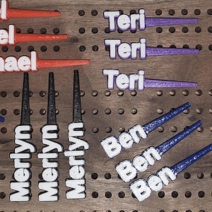 Puede incluir: Una colección de piezas personalizadas para juegos de clavijas con nombres como "Michael", "Damon", "Merlyn", "Ben", "Teri" y "Julie" en letras blancas. Las clavijas son de varios colores, incluyendo rojo, azul, negro, morado y amarillo. Están dispuestas sobre un tablero de clavijas de madera.