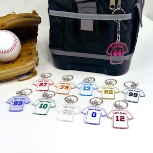 May include: A selection of acrylic jersey-shaped keyrings in various colours and numbers. Each keyring has a silver ring and the text "YOUR NAME" above the number. A baseball, glove, and a black cooler bag are in the background.