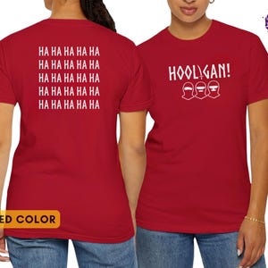 K&ouml;nnte beinhalten: Ein rotes Kurzarm-T-Shirt mit wei&szlig;em Text. Die Vorderseite zeigt das Wort HOOLIGAN! &uuml;ber drei stilisierten Gesichtsmasken. Die R&uuml;ckseite ist mit dem wiederholten Text HA HA HA HA HA in f&uuml;nf Zeilen bedruckt. Das T-Shirt ist von beiden Seiten zu sehen.