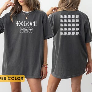 K&ouml;nnte beinhalten: Ein anthrazitfarbenes Kurzarm-T-Shirt mit dem wei&szlig;en Schriftzug HOOLIGAN! und drei Maskensymbolen auf der Vorderseite. Die R&uuml;ckseite des Shirts zeigt den wiederholten wei&szlig;en Text HA HA HA HA HA HA in f&uuml;nf Zeilen. Der Stoff hat eine verwaschene Optik.