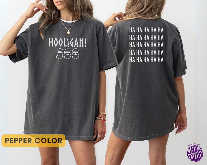 K&ouml;nnte beinhalten: Ein anthrazitfarbenes Kurzarm-T-Shirt mit dem wei&szlig;en Schriftzug HOOLIGAN! und drei Maskensymbolen auf der Vorderseite. Die R&uuml;ckseite des Shirts zeigt den wiederholten wei&szlig;en Text HA HA HA HA HA HA in f&uuml;nf Zeilen. Der Stoff hat eine verwaschene Optik.
