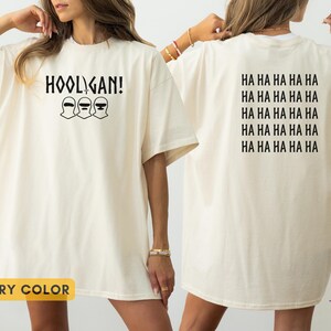 K&ouml;nnte beinhalten: Ein elfenbeinfarbenes T-Shirt mit einer schwarzen Grafik auf der Vorderseite, die den Text HOOLIGAN und drei Sturmhauben-Symbole zeigt. Auf der R&uuml;ckseite ist der Text HA HA HA HA HA f&uuml;nfmal in Schwarz in einem Block aus f&uuml;nf Zeilen abgedruckt.