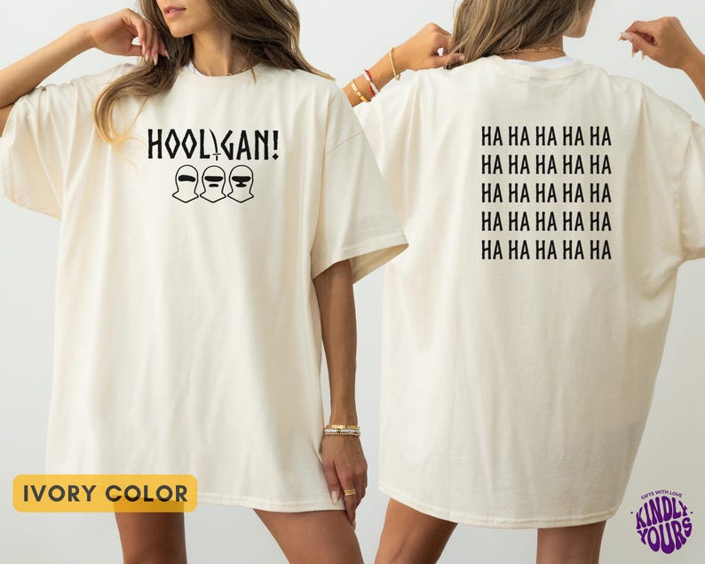 K&ouml;nnte beinhalten: Ein elfenbeinfarbenes T-Shirt mit einer schwarzen Grafik auf der Vorderseite, die den Text HOOLIGAN und drei Sturmhauben-Symbole zeigt. Auf der R&uuml;ckseite ist der Text HA HA HA HA HA f&uuml;nfmal in Schwarz in einem Block aus f&uuml;nf Zeilen abgedruckt.