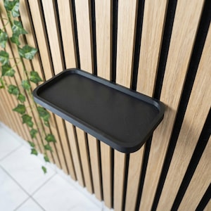 Può includere: Una mensola rettangolare nera montata su una parete a doghe di legno. La mensola, con un bordo leggermente rialzato, è vuota. La parete è composta da doghe di legno verticali. Piante verdi sono visibili a sinistra.