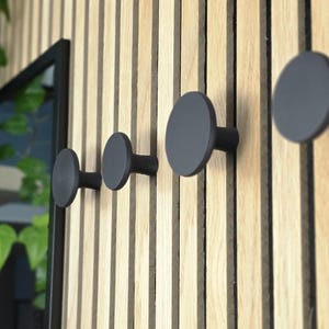 May include: A wooden wall featuring four dark grey, circular coat hooks. The hooks are mounted on vertical wooden slats. The wood has a natural, light brown colour. The hooks are evenly spaced.