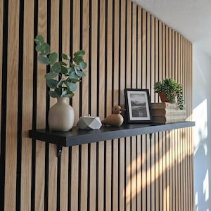 Könnte beinhalten: Ein schwarzes Wandregal an einer vertikalen Holzlattenwand. Das Regal enthält eine beige Keramikvase mit Eukalyptuszweigen, einen grauen geometrischen Kerzenhalter, eine hölzerne Vogelfigur, ein gerahmtes Schwarz-Weiß-Foto und eine Topfpflanze.