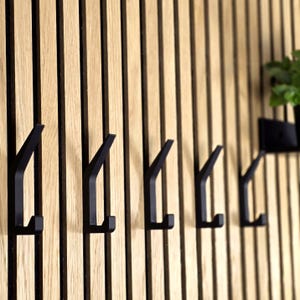 May include: A set of five black metal wall hooks mounted on a vertical wooden slat wall. The hooks have a minimalist, modern design and are evenly spaced. The wood has a natural, light brown color.