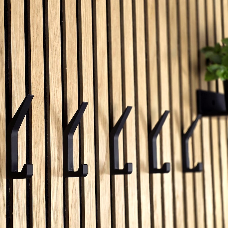 May include: A set of five black metal wall hooks mounted on a vertical wooden slat wall. The hooks have a minimalist, modern design and are evenly spaced. The wood has a natural, light brown color.