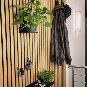 May include: A modern entryway with a wooden slatted wall featuring a black shelf with a potted plant, keys, and sunglasses. A brown leather jacket and a black hat hang on hooks. A second shelf holds a small plant and decorative items.