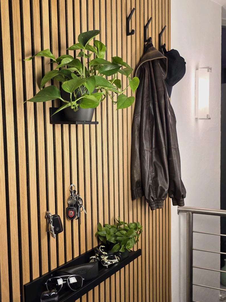 May include: A modern entryway with a wooden slatted wall featuring a black shelf with a potted plant, keys, and sunglasses. A brown leather jacket and a black hat hang on hooks. A second shelf holds a small plant and decorative items.