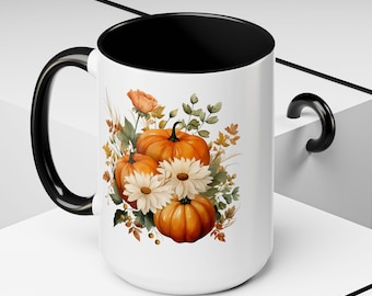 May include: White ceramic mug with a black interior and handle, featuring a watercolor design of pumpkins, daisies, and foliage in shades of orange, white, and green. The mug is a standard size for hot beverages.