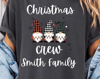 May include: A charcoal gray t-shirt featuring a graphic of three gnomes wearing red, black, and green plaid hats, holding a string of Christmas lights. The text reads "Christmas crew" and "Smith family" in white script and block lettering.