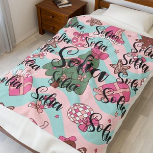 May include: A pink and blue blanket with a repeating pattern of Christmas-themed illustrations, including a Christmas tree, gifts, and gingerbread men. The name "Sofia" is written in a cursive font throughout the design.