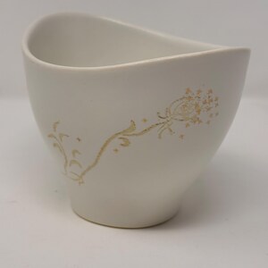 May include: A white ceramic cup with a matte finish and an asymmetrical, curved rim. The exterior features a delicate, gold-colored line drawing of a dandelion seed head with a long, winding stem and small star-shaped accents scattered around.