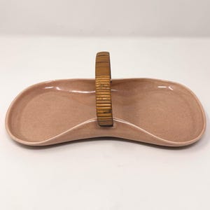May include: A light pink, kidney-shaped ceramic serving dish with a smooth, matte finish. The dish features a central, arched handle wrapped in thin, horizontal strips of natural brown bamboo, creating a distinct contrast with the ceramic base.