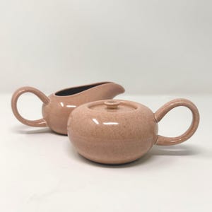 May include: A ceramic gravy boat and a matching lidded sugar bowl in a soft, speckled dusty pink glaze. Both pieces feature rounded bodies and curved handles, with the gravy boat having a wide spout and the sugar bowl a circular lid.