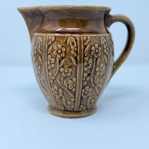May include: A brown glazed ceramic creamer featuring a relief pattern of vertical panels filled with small flowers and leaves. The jug has a curved handle on the right side and a distinct pouring spout on the left, with a smooth rim and base.