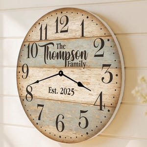 May include: A round wooden wall clock with a distressed, rustic design. The clock face features black numerals, hands, and the text "The Thompson Family Est. 2025". The clock is mounted on a white wall.