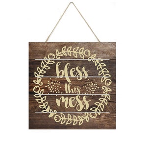 May include: Wooden sign with a rustic brown background and a gold wreath with the text "Bless this mess" in the center.