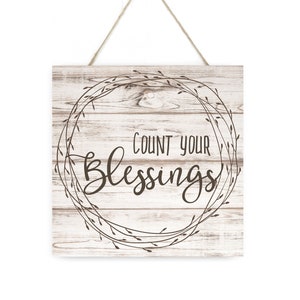 May include: A white wooden sign with a brown wreath and the text "Count your Blessings" in a cursive font.