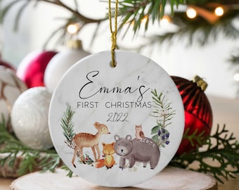 May include: White ceramic Christmas ornament with a marbleized background. The ornament features a deer, fox, bear, and owl in a woodland scene. The text "Emma's First Christmas 2022" is written in black cursive.