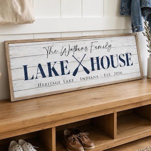 May include: A wooden sign with the text "The Watkins Family LAKE HOUSE" in navy blue, with crossed oars. The sign is on a wooden bench with shoes and a basket. A beige tote bag hangs on the wall.
