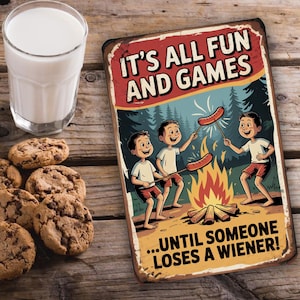 May include: A vintage-style metal sign with the text "IT'S ALL FUN AND GAMES...UNTIL SOMEONE LOSES A WIENER!" features a cartoon illustration of three boys roasting sausages over a campfire. A glass of milk and chocolate chip cookies are on a wooden surface.