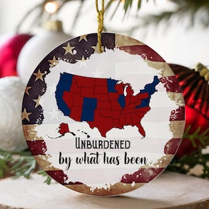 May include: A round, white Christmas ornament with a distressed American flag design. The ornament features a map of the United States with red and blue states. The text "UNBURDENED by what has been" is printed on the ornament.