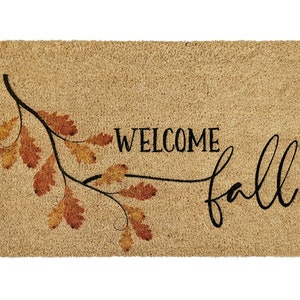 May include: A brown coir doormat with the text "Welcome Fall" in black script. A branch with orange and brown leaves is depicted on the left side of the mat.