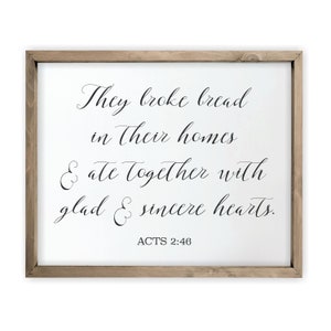 May include: A framed white canvas sign with black calligraphy that reads "They broke bread in their homes & ate together with glad & sincere hearts. ACTS 2:46".