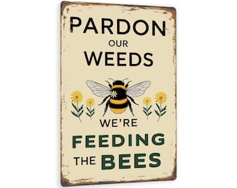 May include: A rectangular metal sign with a distressed finish. The sign features the text "Pardon Our Weeds, We're Feeding the Bees" in black and green. A bumblebee illustration is centered above the text, with yellow flowers on either side.