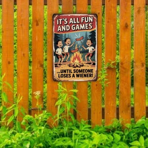 May include: A rectangular metal sign with the text "IT'S ALL FUN AND GAMES" and "...UNTIL SOMEONE LOSES A WIENER!". The sign features a cartoon illustration of three children roasting hot dogs over a campfire. The sign is mounted on a wooden fence.
