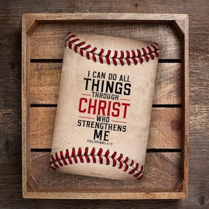 May include: A baseball-shaped sign with a white background and red stitching. The sign features the text "I can do all things through Christ who strengthens me" in black and red lettering. The text "Philippians 4:13" is printed in black below the main text.