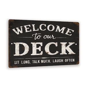 May include: Black and white metal sign with distressed paint. The sign reads "Welcome to our Deck. Sit long, talk much, laugh often."