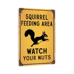 May include: Yellow metal sign with black text and a silhouette of a squirrel. The sign reads "Squirrel Feeding Area Watch Your Nuts".