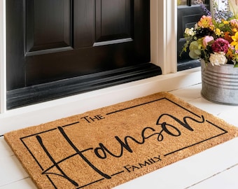 Puede incluir: Una alfombra de coco marrón con el texto "The [family name] Family" en un marco rectangular negro.