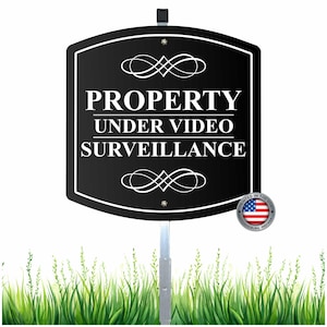 May include: Black property sign with white text that reads "PROPERTY UNDER VIDEO SURVEILLANCE." The sign has decorative flourishes and is mounted on a metal pole. A small emblem with the American flag is in the lower right corner.