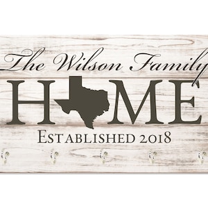 May include: White wooden sign with the text "The Wilson Family HOME Established 2018" and a silhouette of the state of Texas. The sign has five silver hooks.