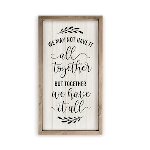 May include: A framed wooden sign with a white background and black text that reads "We may not have it all together but together we have it all". The sign is decorated with small black floral sprigs at the top and bottom.
