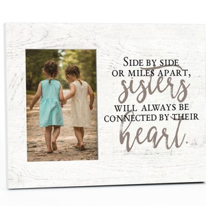 May include: White wood picture frame with a photo of two girls walking hand-in-hand. The frame has a quote that reads: "Side by side or miles apart, sisters will always be connected by their heart."