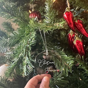 May include: Clear round Christmas ornament with the text "Chateau Ferrari" written in white calligraphy on a green Christmas tree.