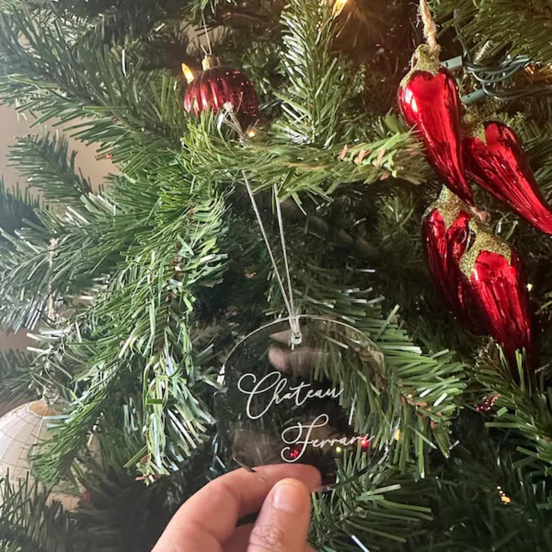 May include: Clear round Christmas ornament with the text "Chateau Ferrari" written in white calligraphy on a green Christmas tree.