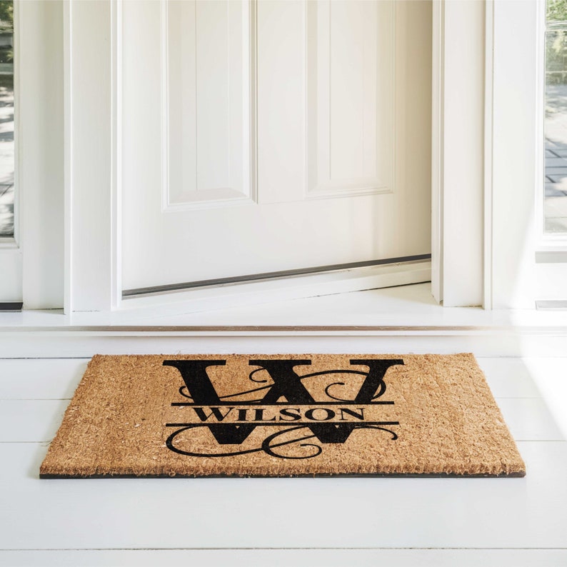 A personalized custom printed coir coconut fiber door mat.  It is printed with a family name and large monogram initial. It is sitting on a white wood porch in front of a beautiful white entry door. The image is bright and sunny and inviting.