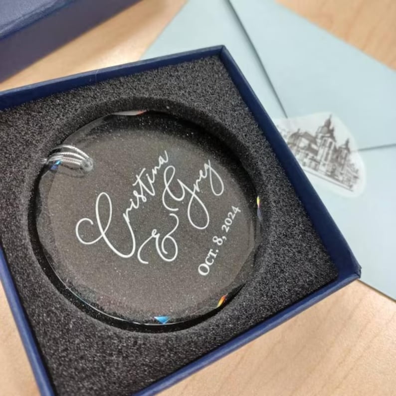 May include: A clear glass ornament with the names "Christina & Greg" and the date "Oct. 8, 2024" engraved in white. The ornament is in a black box with a blue lid.