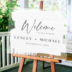 May include: White acrylic wedding welcome sign with black brushstrokes and text that reads "Welcome to our wedding Lesley + Michael December 14, 2024".